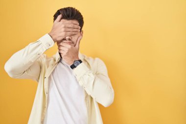 Young hispanic man standing over yellow background covering eyes and mouth with hands, surprised and shocked. hiding emotion 