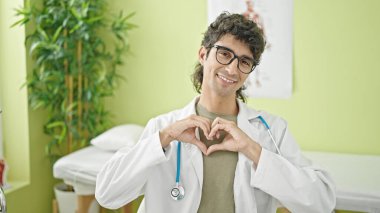 Genç İspanyol doktor gülümsüyor klinikte elleriyle kalp hareketi yapıyor.