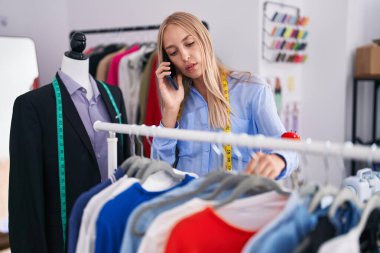 Elbiseleri askıda tutan genç sarışın kadın terzide akıllı telefondan konuşuyor.