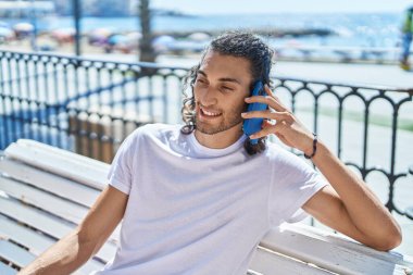 Young hispanic man talking on the smartphone sitting on bench at seaside