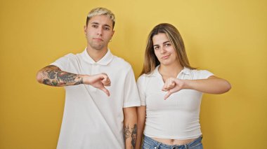 Güzel bir çift yan yana duruyor, ciddi bir yüz ifadesiyle izole edilmiş sarı arka planda baş parmak hareketi yapıyorlar.