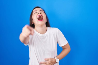 Orta yaşlı Latin bir kadın mavi arka planda durup sana gülüyor, eli vücudunun üzerinde kamerayı işaret ediyor, utanç ifadesi. 