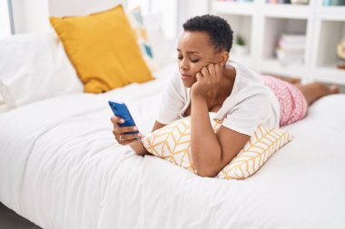 Afrika kökenli Amerikalı kadın yatak odasında endişeli bir ifadeyle akıllı telefon kullanıyor.