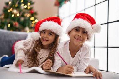 Erkek ve kız kardeş Noel Baba mektubu yazıyorlar. Evdeki Noel ağacının yanında kanepeye uzanmışlar.