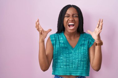 Genç Afro-Amerikan örgüleri pembe arka planda çılgın ve kızgın bir şekilde dikilmiş agresif bir ifadeyle ve kollarıyla bağırıyor. hüsran kavramı. 