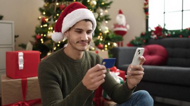 Genç İspanyol adam akıllı telefon ve kredi kartı kullanıyor. Evdeki Noel ağacının yanında yerde oturuyor.