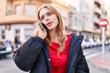 Young blonde woman talking on smartphone with serious expression at street
