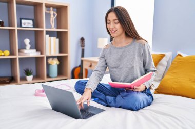 Young beautiful hispanic woman student reading book studying on bed at bedroom