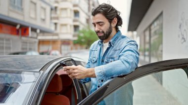 Genç İspanyol adam akıllı telefon kullanıyor. Sokakta arabanın yanına yaslanıyor.