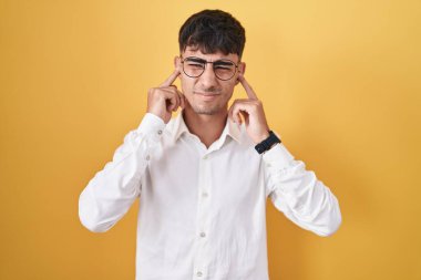 Sarı arka planda dikilen İspanyol genç adam yüksek sesli müziğin gürültüsü için parmaklarıyla kulaklarını kapatıyor. sağır kavramı. 