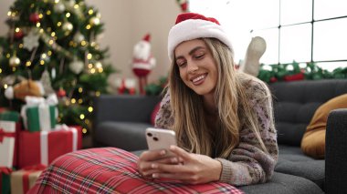 Evdeki Noel ağacının yanındaki koltukta akıllı telefon kullanan genç sarışın kadın.