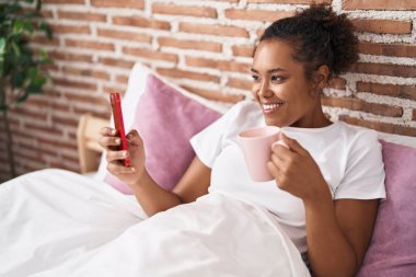 African american woman using smartphone drinking coffee at bedroom