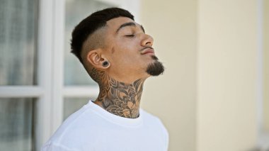 Genç Latin bir adam dinleniyor, açık havada temiz hava soluyor, güneşli bir şehir sokağında gözleri kapalı, rahatlamış ve sağlığını dengeliyor.