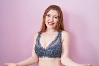 Pembe arka planda iç çamaşırı giyen kızıl saçlı kadın güler yüzlü ve kolları açık bir şekilde hoş geldin diyor, olumlu ve kendinden emin selamlar. 