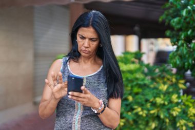 Orta yaşlı İspanyol kadın sokakta ciddi bir ifadeyle akıllı telefon kullanıyor.