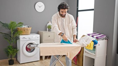 Young hispanic man listening to music ironing clothes at laundry room