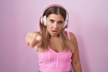 Young blonde woman listening to music using headphones pointing displeased and frustrated to the camera, angry and furious with you 