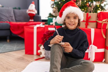 Adorable hispanic boy writing santa claus letter sitting by christmas tree at home