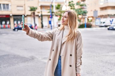 Young blonde woman smiling confident making selfie by the smartphone at street