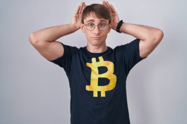 Beyaz, sarışın, bitcoin tişörtü giyen tavşan kulağı jestleri yapan elleri kuşkucu ve alaycı görünen bir adam. Paskalya tavşanı konsepti. 