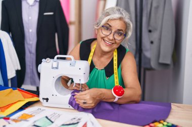 Orta yaşlı gri saçlı kadın terzi dükkanında dikiş makinesi kullandığından emin bir şekilde gülümsüyor.