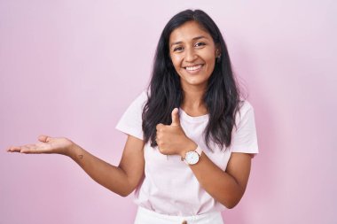 Pembe arka planda duran genç İspanyol kadın avuç içini gösteriyor ve baş parmaklarıyla iyi bir hareket yapıyor, mutlu ve neşeli gülümsüyor. 