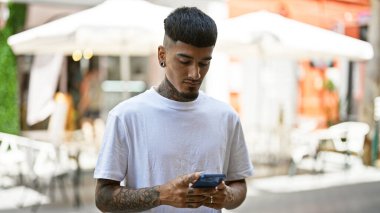 Havalı Latin adam duruyor, ciddi ifade ediyor, şehir kafeteryasının terasında güneşli şehir semalarında akıllı telefondan yazı yazıyor.