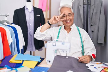 Orta yaşlı bir adam, gri saç terzisi dikiş makinesi kullanıyor. Yüzünde parmakları olan barış sembolü yapıyor. Neşeli bir şekilde zafer işareti yapıyor. 