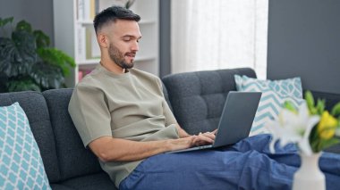 Genç İspanyol adam evdeki koltukta dizüstü bilgisayar kullanıyor.