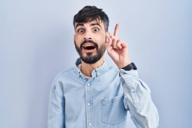 Mavi arka planda duran sakallı genç İspanyol adam başarılı bir fikir işaret ediyor. Heyecanlı ve mutlu. Bir numara.. 