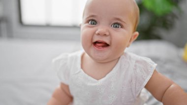 Sevimli yeni uyanmış bebek rahatlamış, yatakta oturmuş, rahat yatak odasında rahatça gülümsüyor.