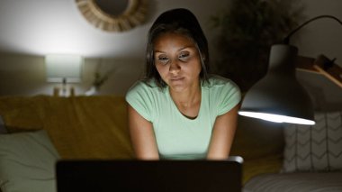 Evdeki kanepede dizüstü bilgisayar kullanan genç Latin kadın.