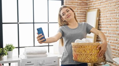 Genç sarışın kadın çamaşır odasında çamaşır sepetiyle selfie çekiyor.