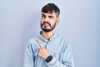 Mavi arka planda duran sakallı genç İspanyol adam endişeli, endişeli ve şaşırmış ifadesini işaret ediyor. 