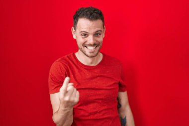 İspanyol asıllı genç adam kırmızı arka planda duruyor. Buraya el kol hareketi yaparak mutlu bir şekilde karşılamaya ve gülümsemeye davet ediyor. 
