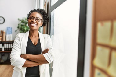 Afro-Amerikalı iş kadını gülümseyen, kendine güvenen, kollarını kavuşturup ofiste el kol hareketi yapan.