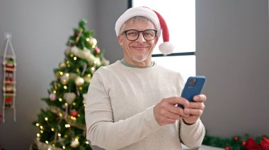 Orta yaşlı, kır saçlı, akıllı telefon kullanan bir adam Noel ağacının yanında duruyor.