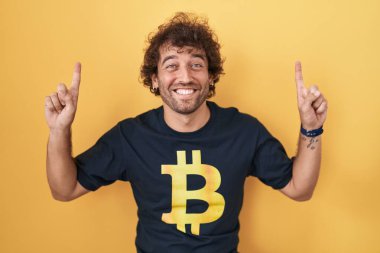 İspanyol asıllı genç adam Bitcoin t-shirt giyiyordu. Şaşırmış, şaşırmış, parmaklarıyla işaret ediyor ve kollarını kaldırıyordu.. 