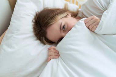 Young caucasian woman covering face with blanket lying on bed at bedroom