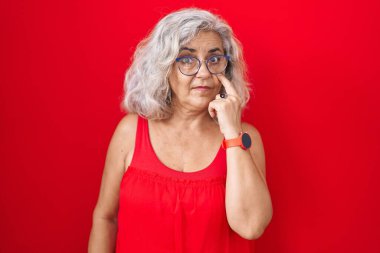 Kırmızı arka planda gri saçlı orta yaşlı bir kadın. Gözünü dikip jestini izliyor. Şüpheli bir ifade. 