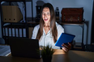 Young brunette woman working at the office at night winking looking at the camera with sexy expression, cheerful and happy face. 