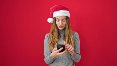 Kafkasyalı genç bir kadın noel şapkası takıyor ve izole edilmiş kırmızı arka plan üzerinde akıllı telefon kullanıyor.