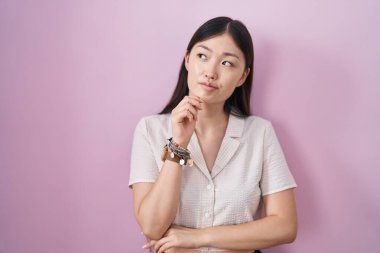 Çinli genç bir kadın pembe arka planda, elleri çenesinde soru sorarken, düşünceli bir ifade takınıyor. Düşünceli bir yüzle gülümsüyor. şüphe kavramı. 