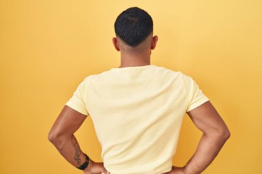 Young hispanic man standing over yellow background standing backwards looking away with arms on body 