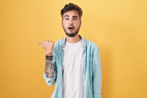 Young hispanic man with tattoos standing over yellow background surprised pointing with hand finger to the side, open mouth amazed expression. 