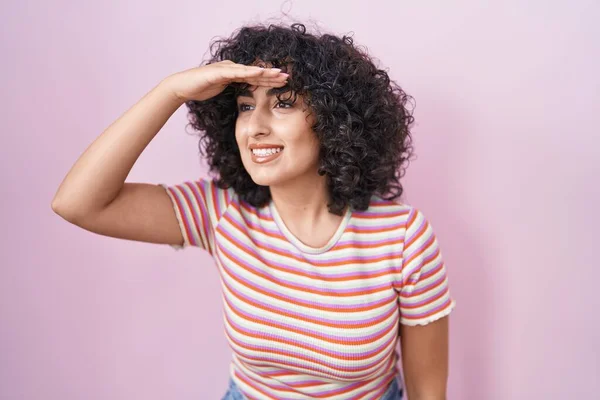 Young middle east woman standing over pink background very happy and smiling looking far away with hand over head. searching concept.