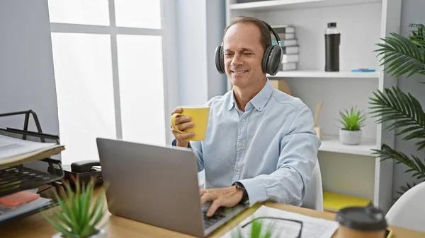 Cheerful middle age man, a successful executive, joyfully working at his office desk, listening to music and sipping his morning espresso coffee, brightening up the indoor workplace.