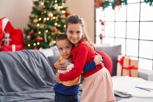 Brother and sister hugging each other standing by christmas tree at home