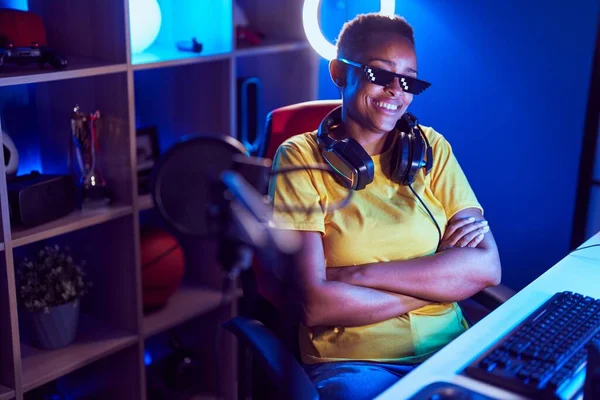 African american woman streamer wearing thug life glasses sitting with arms crossed gesture at gaming room