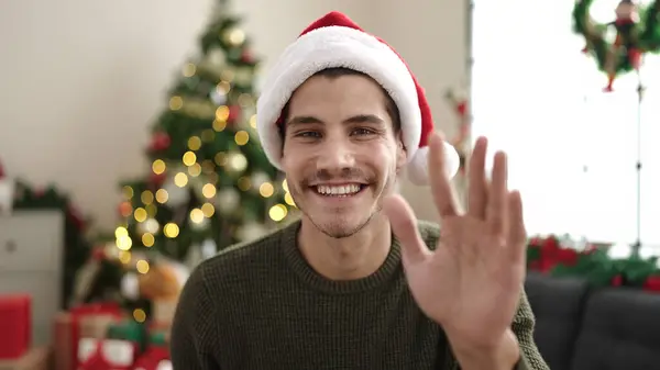 Young hispanic man saying hello with hand sitting on floor by christmas tree at home